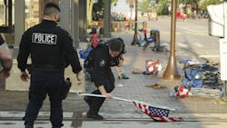A police officer picks up a water-logged American flag Tuesday, left behind after the mass shooting during a Fourth of July parade in Highland Park, IL. A police officer picks up a water-logged American flag Tuesday, left behind after the mass shooting during a Fourth of July parade in Highland Park, IL.