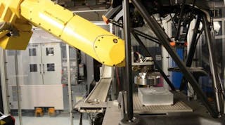 Robot places a machined bearing into the Equator&rsquo;s measuring envelope (foreground), with part conveyor and Okuma twin-spindle lathe in background. Aluminum block on Equator&rsquo;s fixture plate has hole in center which Equator uses to set its coordinate system. Part is placed in center of block for measurement.